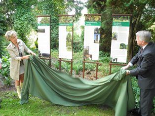 Een vrouw (links) en een man (rechts) houden een groene doek vast, ze staan lachend bij vier banners aan een groene plantenborder in Arboretum Kalmthout, onthulling van infoborden bij de boomplanting.