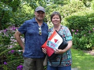 Een man en een vrouw poseren lachend naast bloeiende borders in de tuin van Arboretum Kalmthout. De vrouw houdt een schoudertas vast en toont een ticket waar Terra Nova op staat.