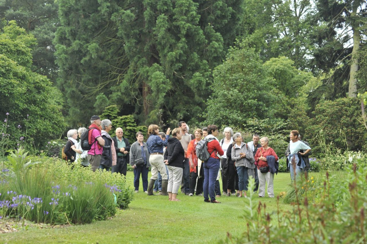 Een groep bezoekers luistert naar de gids in Arboretum Kalmthout, op het gazon omgeven door bloemborders en grote bomen. Licht bewolkte dag in juni.
