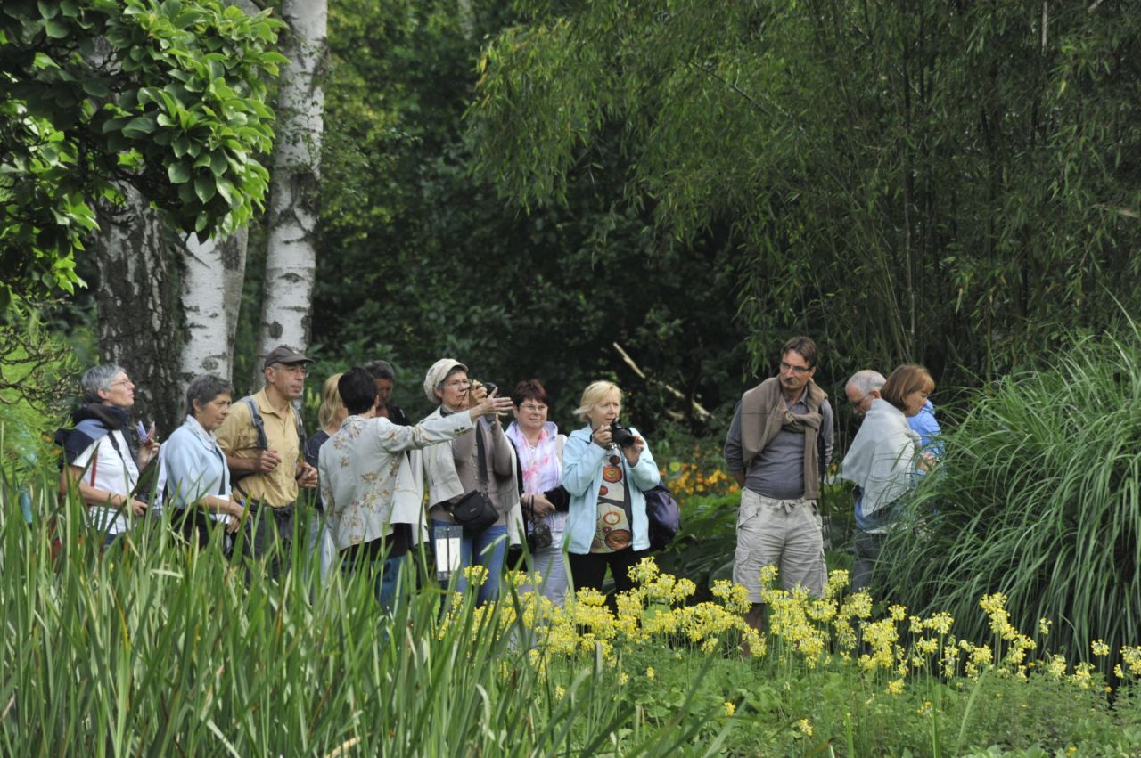 Een gidst loopt op kop van een rij geblinddoekte bezoekers. Twee mannen, twee vrouwen en twee kinderen houden zich vast aan het touw waarmee de gids hen meeneemt doorheen de arboretumtuin. Deze zintuiglijke wandeling heet Op Goed Gevoel.