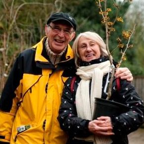 Winter in Arboretum Kalmthout. Twee bezoekers, een ouder koppel,  poseren in het plantencentrum. De vrouw draagt een Hamamelis in pot, de man legt zijn arm om haar schouder. Ze lachen.