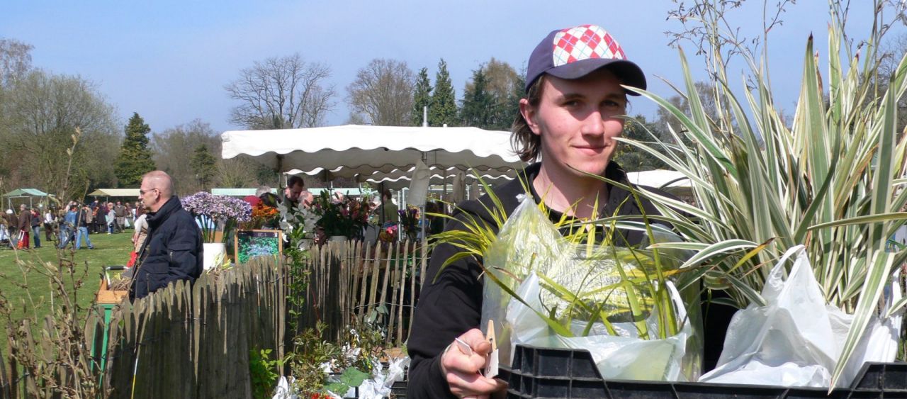 Zonnige Plantendag in Arboretum Kalmthout, zicht op de plantenweide met kraampjes en de plantenparking met een rij verkochte planten. Een jonge tuinier of student draagt een plastieken bak met planten en kijkt in de lens.