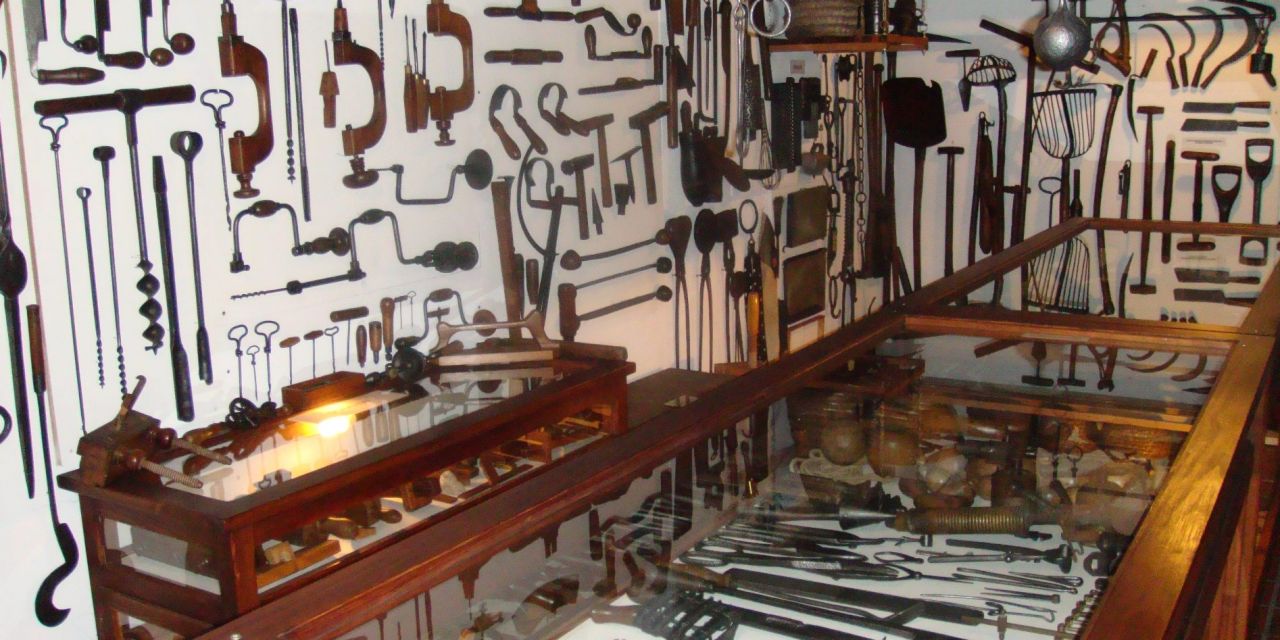 Binnenzicht in het heemkundig museum. Aan de muur en in de houten vitrinetafels een verzameling houten en metalen werktuigen zoals handboren, zagen en tangen.