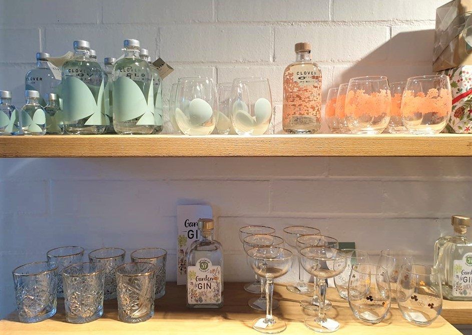 Feestelijke glazen en flessen gin van het Kalmthoutse merk Cover Gin staan uitgestald op twee houten wandplanken in de cadeau- & boekenshop van Arboretum Kalmthout.