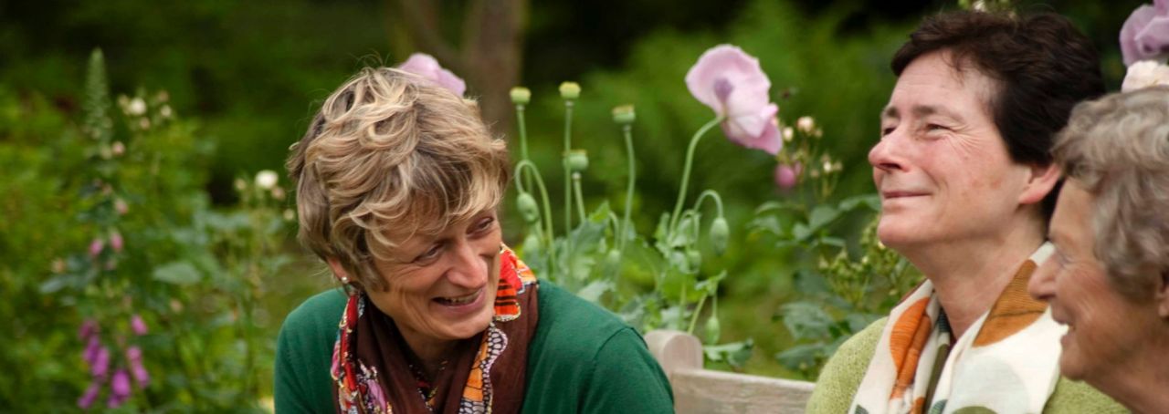 Op een houten tuinbank zitten drie dames. Ze lachen en drinken een glaasje wijn. Op de achtergrond hoge, groene borderplanten als vingerhoedskruid en zachtroze papaver.