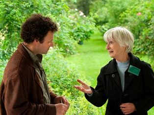 Een dame praat met een man, ze is gids in de plantentuin. Ze draagt een naambordje en geeft uitleg. Op de achtergrond frisgroene stuiken, gescheiden door een graspad.