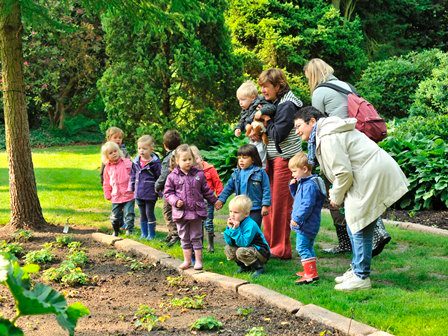 Arboretum Kalmthout. Een gids en twee kleuterjuffrouwen staan bij een groepje kleuters en wijzen hen op iets. De kinderen geven elkaar een hand en kijken om zich heen.