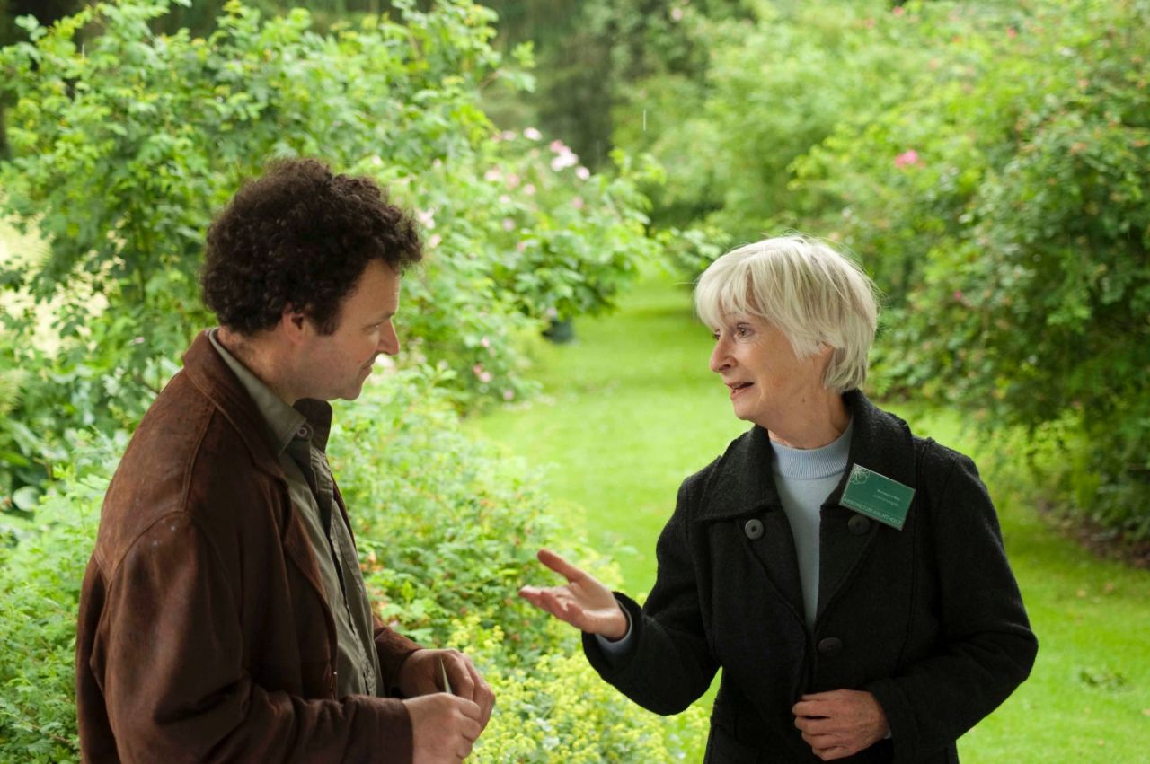 Een dame praat met een man, ze is gids in de plantentuin. Ze draagt een naambordje en geeft uitleg. Op de achtergrond frisgroene stuiken, gescheiden door een graspad.