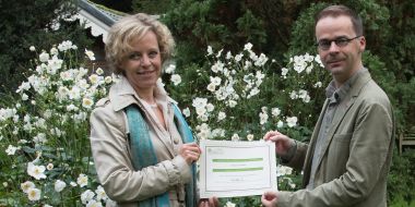 Gedeputeerde van de provincie Antwerpen Inga Verhaert houdt samen met conservator-directeur Abraham Rammeloo het ArbNet certificaat vast, in de tuin van Arboretum Kalmthout. Op de achtergrond bloeien witte herfstanemonen uitbundig in de borders. 
