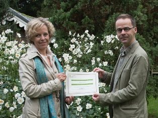 Gedeputeerde van de provincie Antwerpen Inga Verhaert houdt samen met conservator-directeur Abraham Rammeloo het ArbNet certificaat vast, in de tuin van Arboretum Kalmthout. Op de achtergrond bloeien witte herfstanemonen uitbundig in de borders.