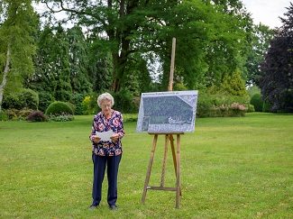 Een oudere dame geeft een toespraak bij een plattegrond op een staander op het gazon van een grote tuin met rijkelijke borders en oude bomen op de achtergrond.