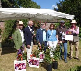 Vijf mannen en een vrouw poseren op een zonnige dag in een tuin voor een aantal rozenstruiken in pot, een van hen houdt fier een certificaat voor met een afbeelding van een roos, doop van een roos tijdens de Tuin- en Plantendagen van Hex 2017.