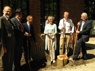 Een plechtig moment bij een boomplanting in Arboretum Kalmthout, 5 mannen en 1 vrouw poseren met een schop in de hand voor de gevel van een gebouw en bij een net aangeplante stek van een druivelaar.