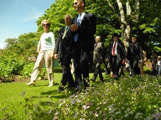 Een groep gasten maakt een wandeling door de zonnige tuin van Arboretum Kalmthout, onder andere de ambassadeur van Slovenië en de burgemeester van de Sloveense stad Maribor. Op de voorgrond groene borders met bloemetjes. 
