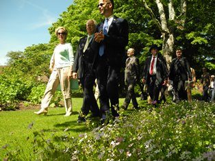 Een groep gasten maakt een wandeling door de zonnige tuin van Arboretum Kalmthout, onder andere de ambassadeur van Slovenië en de burgemeester van de Sloveense stad Maribor. Op de voorgrond groene borders met bloemetjes.