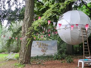 Bolvormige witte tent, zwevend tussen de bomen in Arboretum Kalmthout. Een houten ladder leunt tegen de bol aan, er hangen feestslingers op en er staan tuinstoelen, een klein tafeltje en een banner van Terra Nova, de zomercampagne van de provincie Antwerpen.