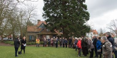 Een cirkel mensen staat op een grasveld rond twee personen die een toespraak houden tijdens de inhuldiging van de tuin achter de cottage in Arboretum Kalmthout. Op de achtergrond een cottage en een monumentale, oude conifeer.