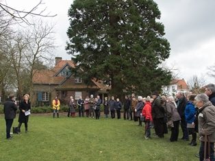 Een cirkel mensen staat op een grasveld rond twee personen die een toespraak houden tijdens de inhuldiging van de tuin achter de cottage in Arboretum Kalmthout. Op de achtergrond een cottage en een monumentale, oude conifeer.