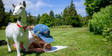 Een meisje zit op de knieën op een groot grasveld en vult een kaartje in, een kunststoffen geitje staat naast haar, zonnige zomerdag in Arboretum Kalmthout